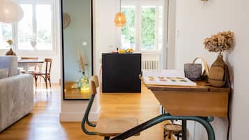 Ein gemütliches Wohnzimmer mit einer Couch, einem Holzesstisch und Stühlen. Es gibt eine schwarze Kommode mit Dekorationsgegenständen und ein Fenster mit Blick auf Grünflächen.