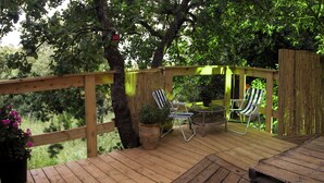 Una terraza de madera con dos sillas a rayas y una pequeña mesa, rodeada de árboles.