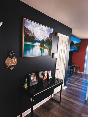 A modern interior with a black wall, a large framed landscape painting, a white door, and a flat-screen TV mounted above it. - 2BR · High Point · Hickswood Townhome (High Point)