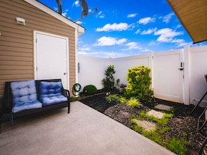 A residential backyard with a covered patio, a white garage door, a white fence, and a garden with various plants. - 2BR · High Point · Hickswood Townhome (High Point)