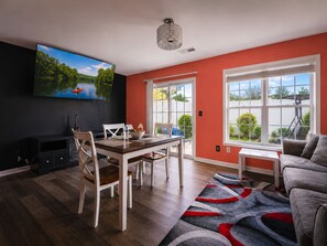 A modern living room with a dining area, a flat-screen TV mounted on the wall, a large window with a view of greenery, and a grey sofa. - 2BR · High Point · Hickswood Townhome (High Point)