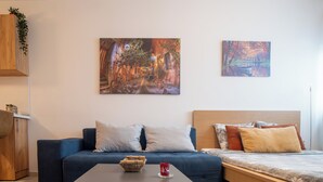 Ein modernes Wohnzimmer mit einer Couch, einem Couchtisch und einem Bett mit Holzvorhang. An der Wand hängen zwei Gemälde, ein Holzschrank mit einer Pflanze und ein kleiner Tisch mit einem Korb und einer roten Tasse.