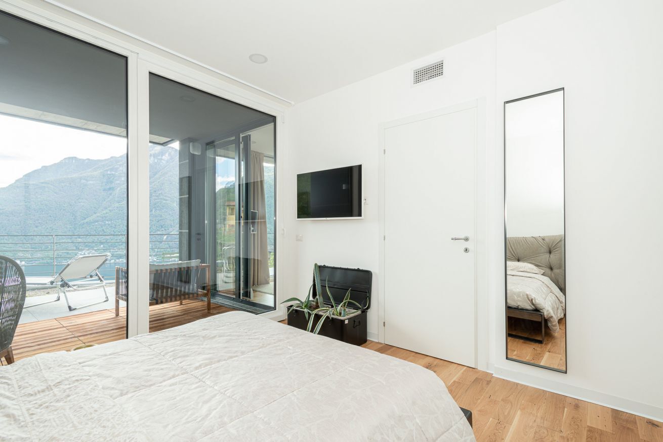 A bedroom with a large bed, a sliding glass door leading to a balcony, a mounted TV, and a mirror on the wall.