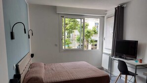 A bedroom with a bed, a desk with a computer and TV, a window with blinds, and a view of buildings and trees outside. - Gloria Maris Apartment (Menton)