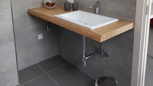 A modern bathroom with a wooden countertop, white sink, and a large mirror. There are two hair dryers mounted on the wall, a trash can, and a small bowl of fruit on the countertop. - Gloria Maris Apartment (Menton)