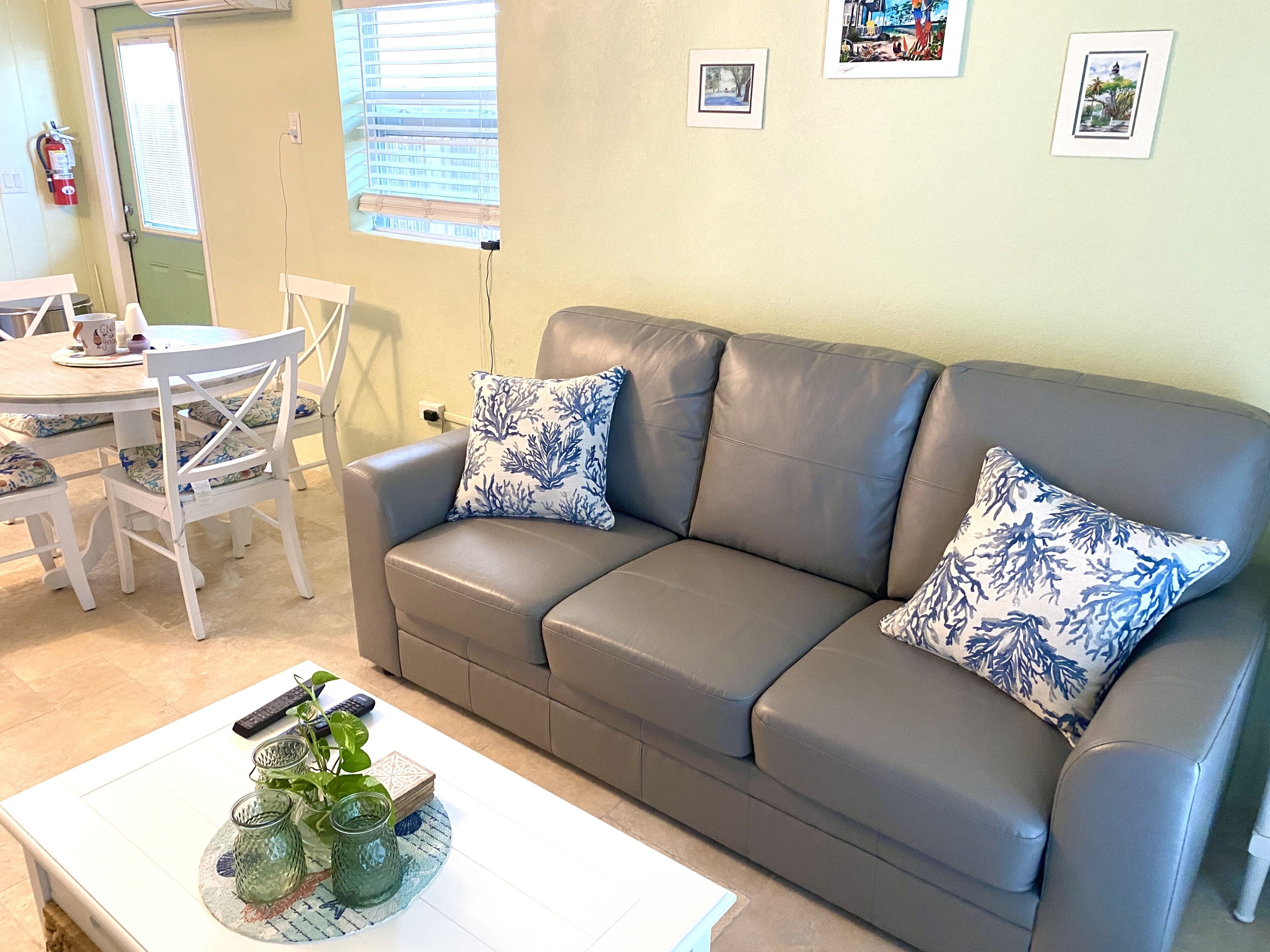 Ein Wohnzimmer mit grauem Sofa, zwei blauen Kissen mit Muster, einem weißen Couchtisch mit Dekoration, einem Esstisch mit Stühlen und einem Fenster mit Jalousien.