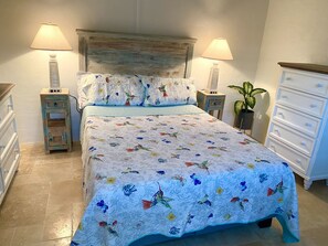 Une chambre à coucher avec une couvre-lit à motifs floraux, deux lampes de chevet, une tête de lit en bois, une table de chevet avec une lampe, une commode et une plante en pot.