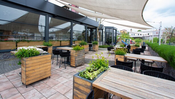 An outdoor patio with wooden tables and chairs, planters with greenery, and a large canopy providing shade. - Hotel Grand (Banja Luka)