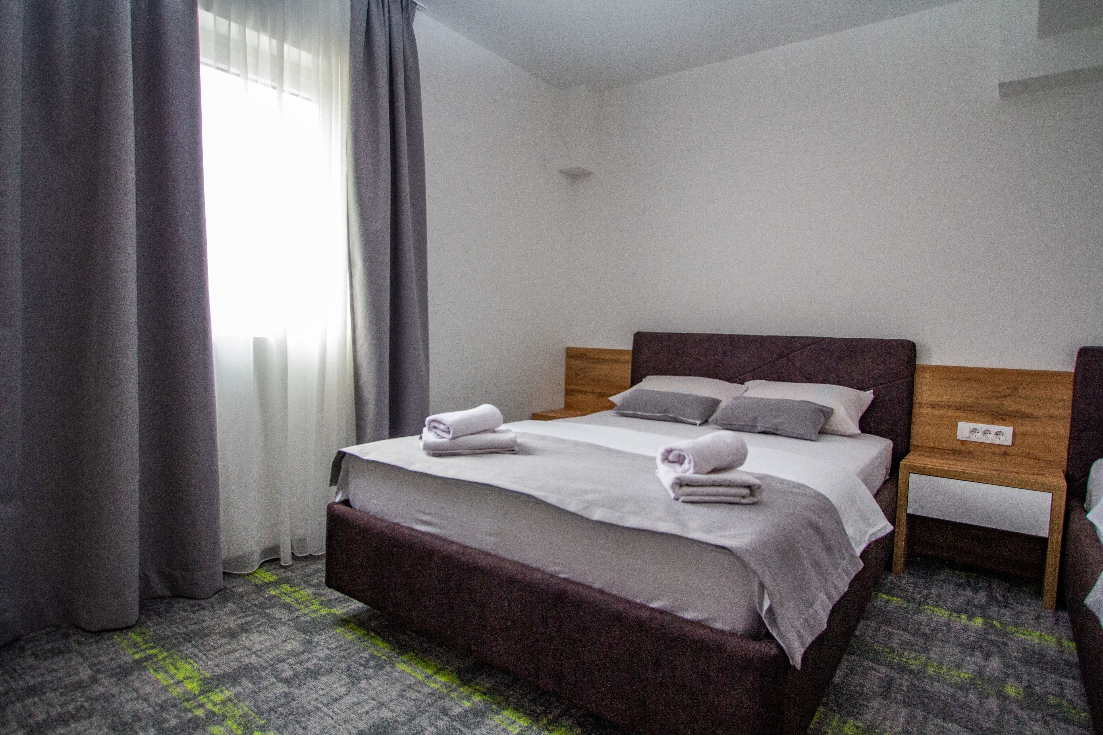 Quarto de hotel com uma cama grande, cabeceira de madeira, mesa de cabeceira e janela com cortinas.