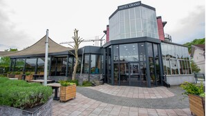 A modern hotel with a glass facade, outdoor seating, and a covered patio area. - Hotel Grand (Banja Luka)