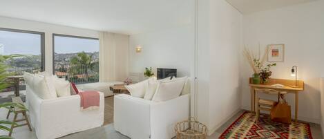 A modern living room with white sofas, a TV, and a large window offering a city view.