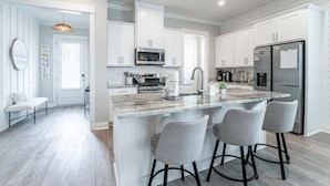 A modern kitchen with white cabinets, a central island with bar stools, stainless steel appliances, and a view into the adjacent dining area. - Luxury 4BR/4BA Beach Retreat Game Room, Resort Pool, & Prime Location (Orange Beach)