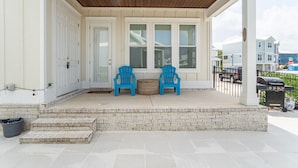 A covered porch with two blue chairs, a grill, and a view of the street. - Luxury 4BR/4BA Beach Retreat Game Room, Resort Pool, & Prime Location (Orange Beach)