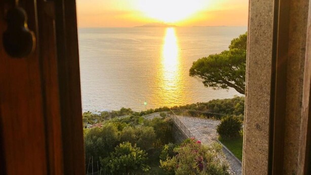 Blick auf das Meer im Sonnenuntergang durch eine offene Tür, im Vordergrund ein Steinweg und Grünpflanzen.