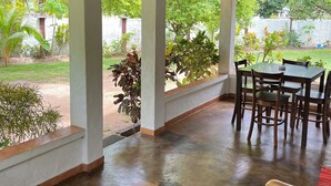 Un espace extérieur abrité, doté d’une table et de chaises en bois, d’un banc et de grandes fenêtres donnant sur un jardin.