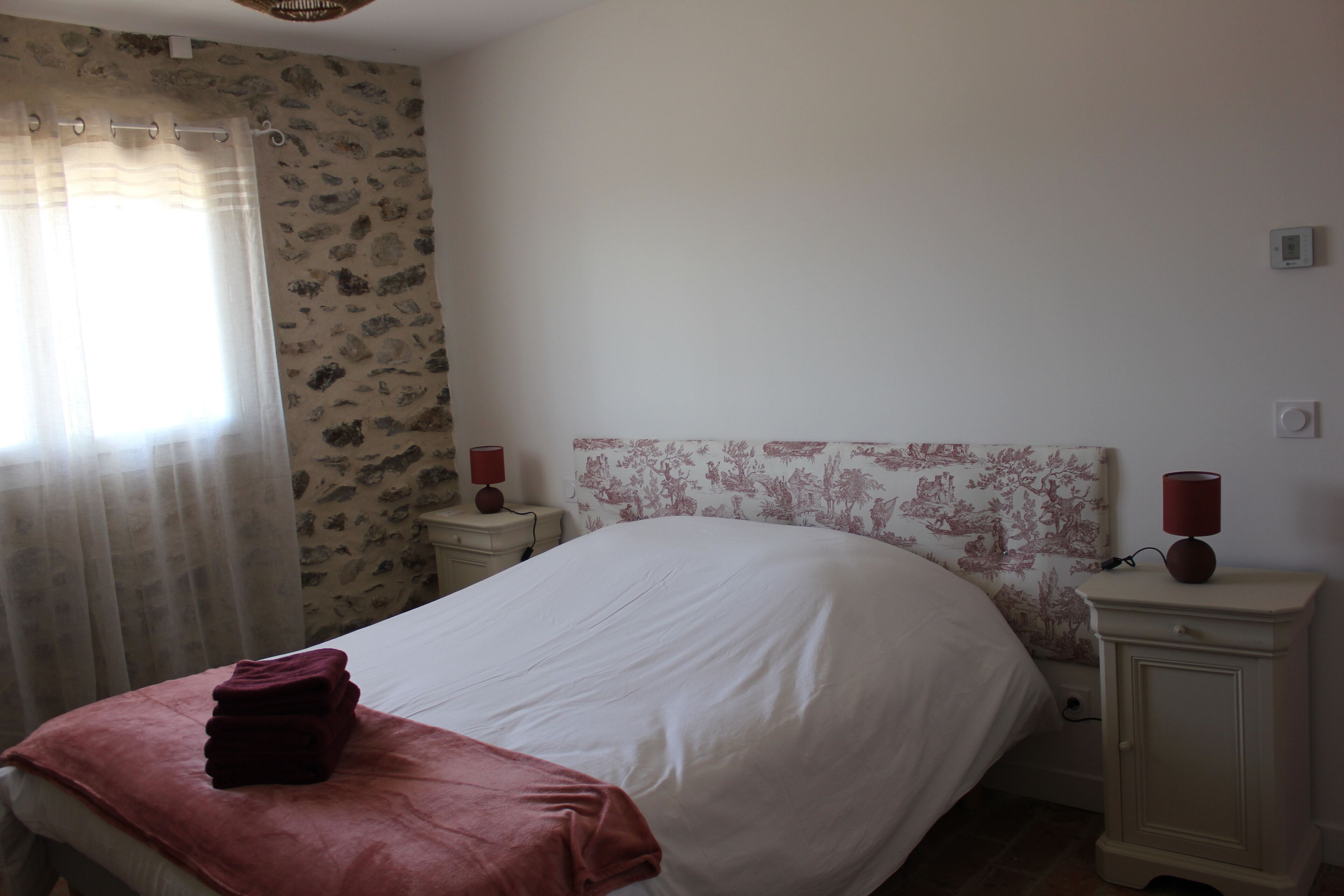 Une chambre à coucher avec un lit, une table de chevet avec une lampe, une fenêtre avec des rideaux et un mur orné d’un motif floral.