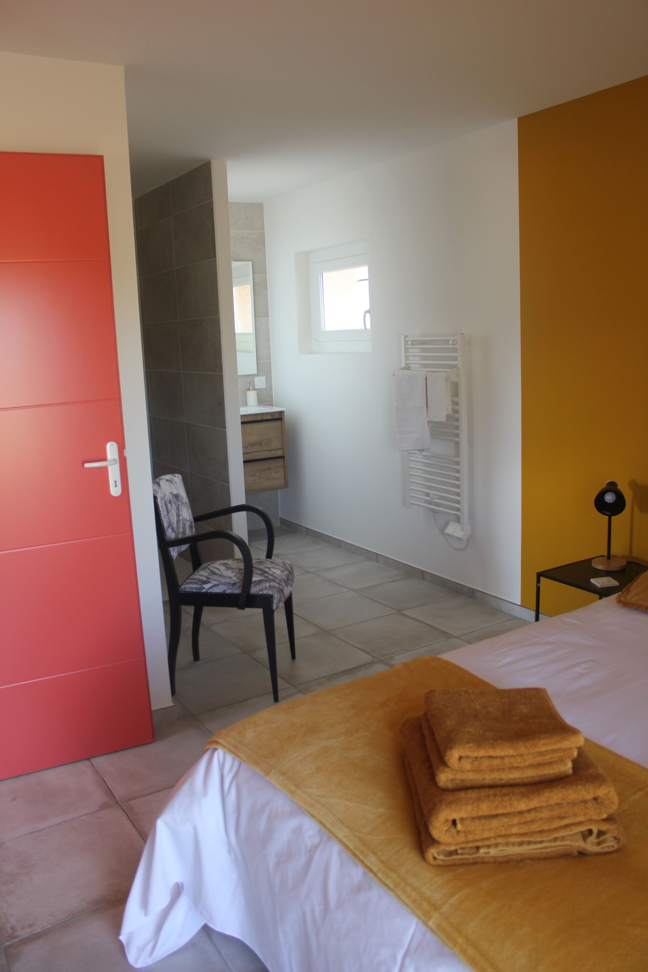 A hotel room with a bed, a chair, a red door, a white wall, a mirror, a towel rack, and a small table with a lamp.