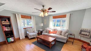 A living room with a wooden floor, a ceiling fan, a sofa, a coffee table, a bookshelf, and a window with curtains.
