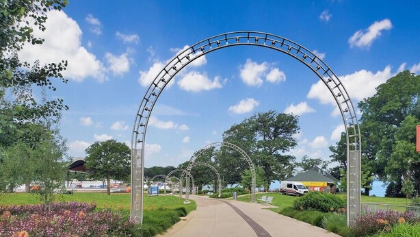 A pathway leading through a park with a metal archway, surrounded by greenery and flowers.