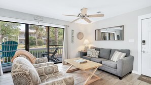 Ein Wohnzimmer mit grauem Sofa, gemusterten Sesseln, einem Holztisch, einem Deckenventilator und einem großen Fenster mit Blick auf einen Balkon und viel Grünzeug.