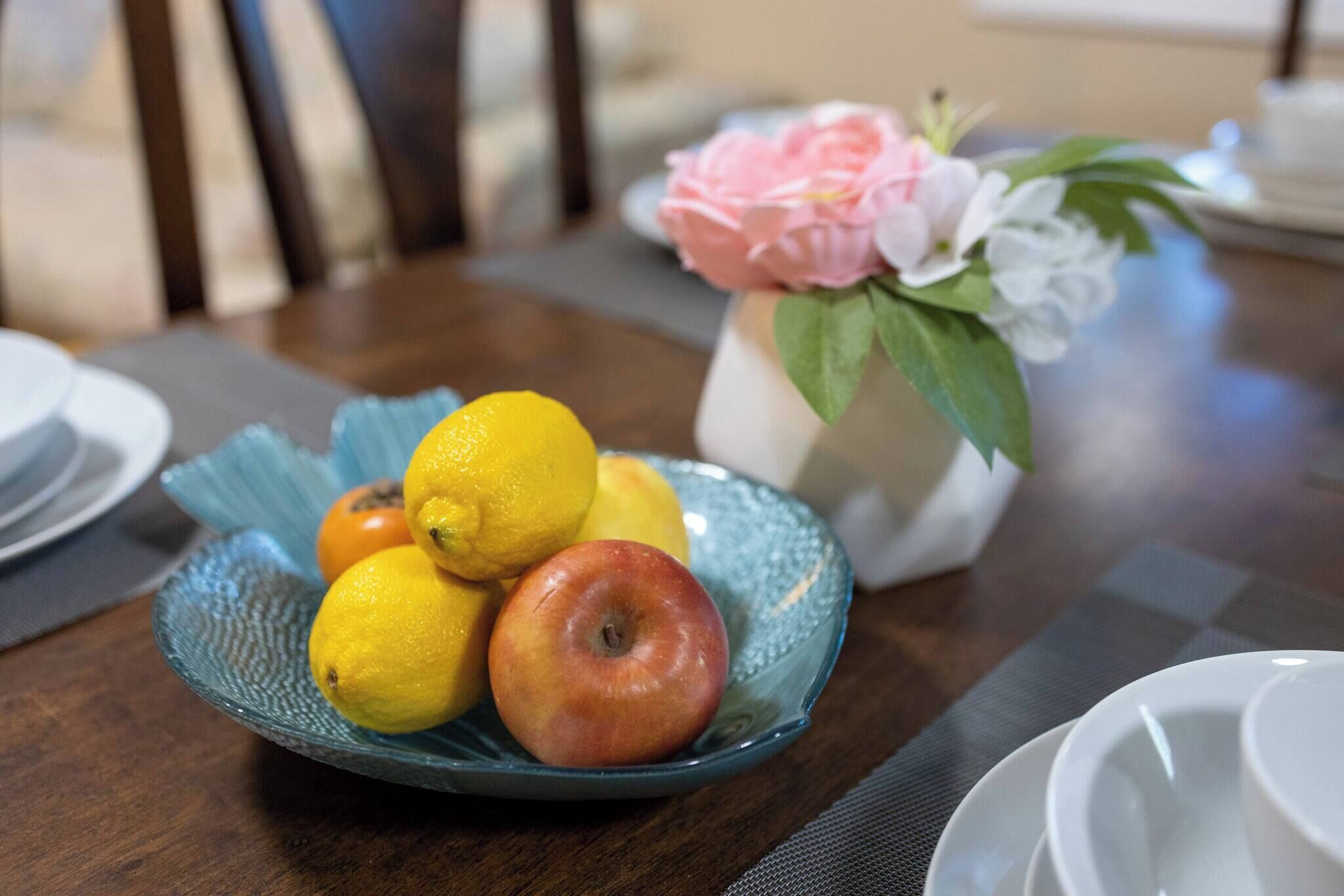 Una mesa con un cuenco de vidrio azul lleno de frutas, un florero con flores y un mantel a cuadros.
