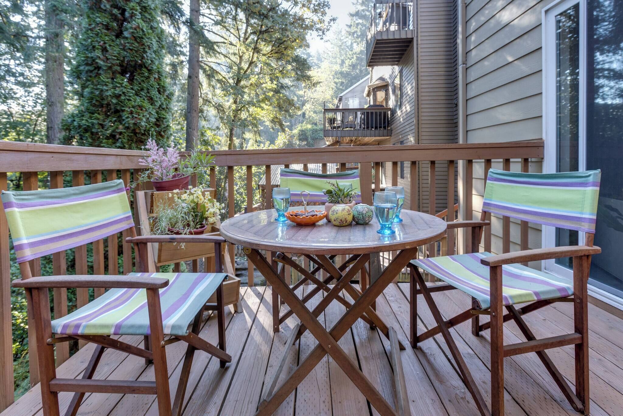 A wooden deck with a round table and striped chairs, surrounded by potted plants and a view of a building.