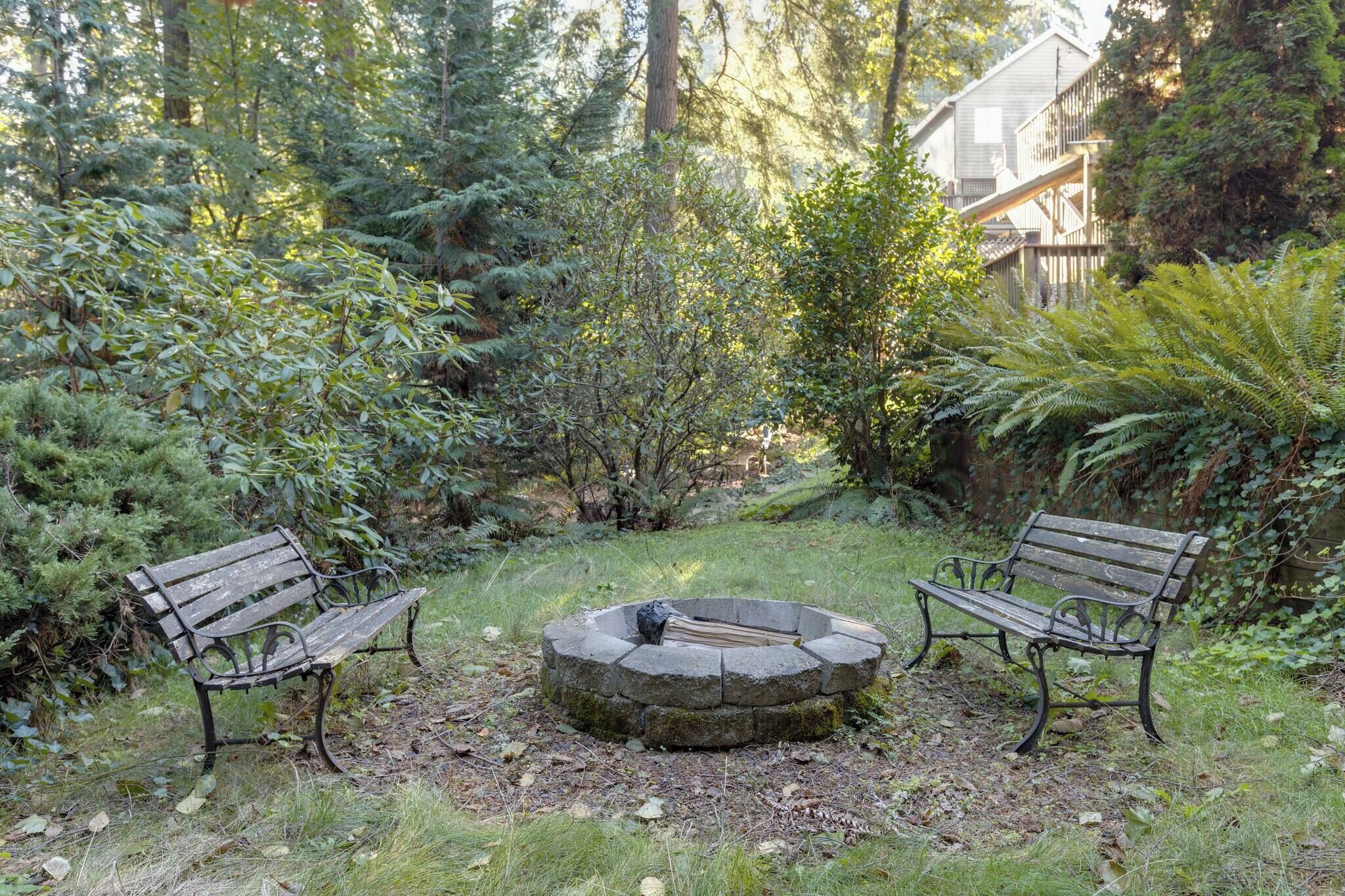 A circular stone fire pit with two benches on either side, surrounded by dense greenery and trees.