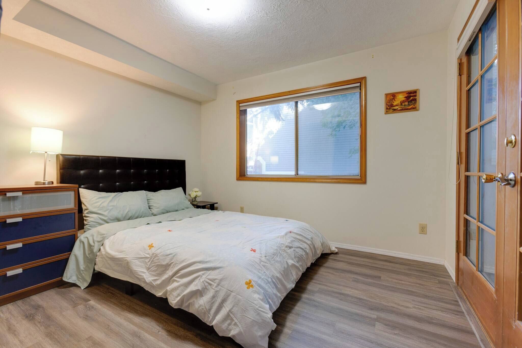Ein Schlafzimmer mit Bett, Kommode, Nachttisch, Lampe, Fenster und Tür.