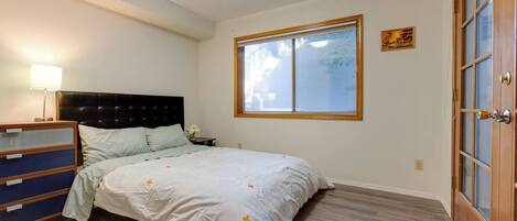 Ein Schlafzimmer mit Bett, Kommode, Nachttisch, Lampe, Fenster und Tür.