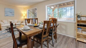 A dining area with a wooden table set for four, surrounded by chairs, with a view of a deck and trees through large windows. - Oregon Jade Lodging- Spacious 2 Bedrooms, Ev Charger, Quite By Stream (West Linn)