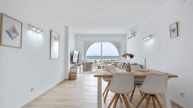 Un comedor moderno con una mesa de madera, sillas blancas y vistas al mar a través de grandes ventanales.