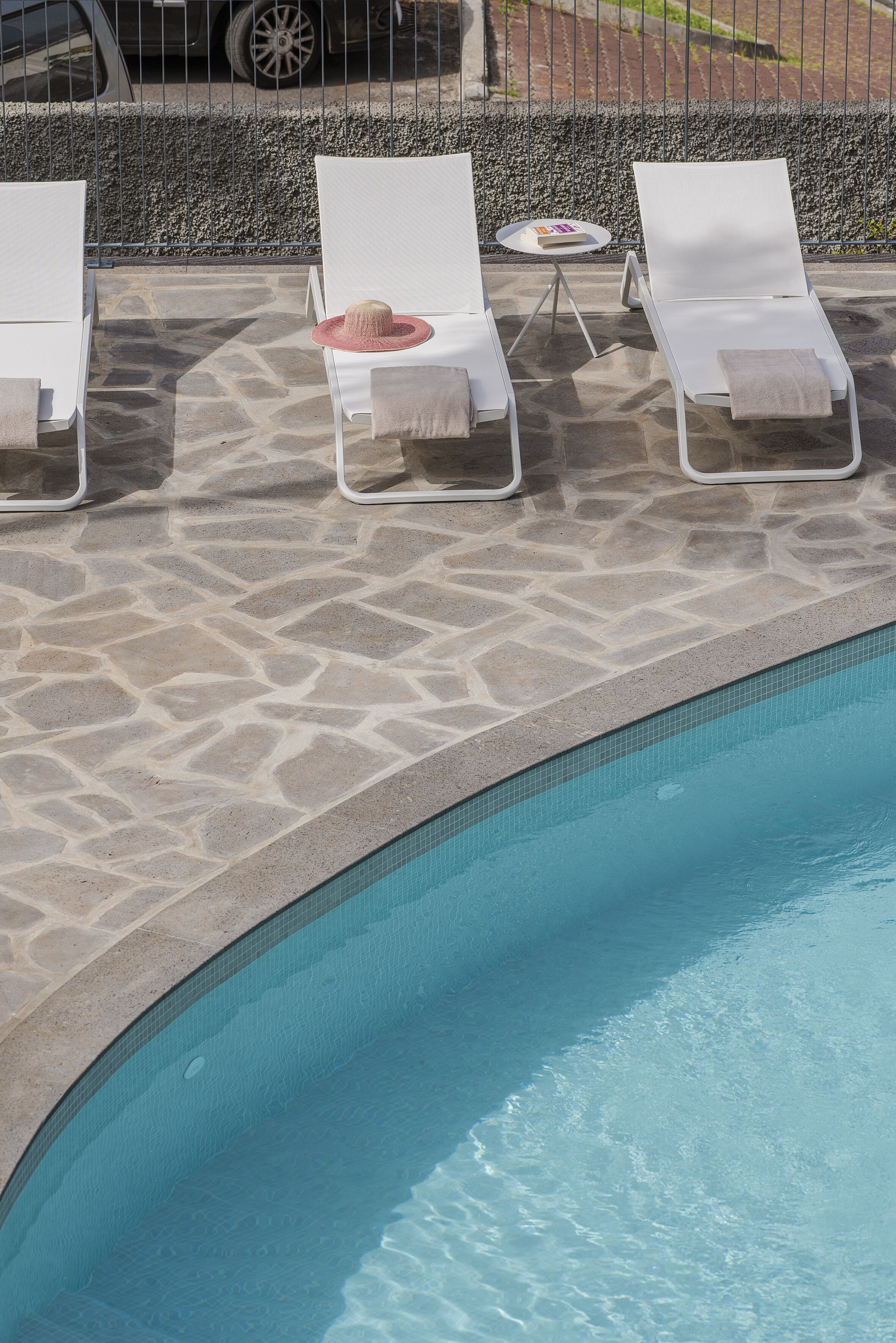 Un espace aménagé au bord de la piscine, avec trois chaises longues blanches, une petite table ronde et un chapeau posé sur l’une des chaises.