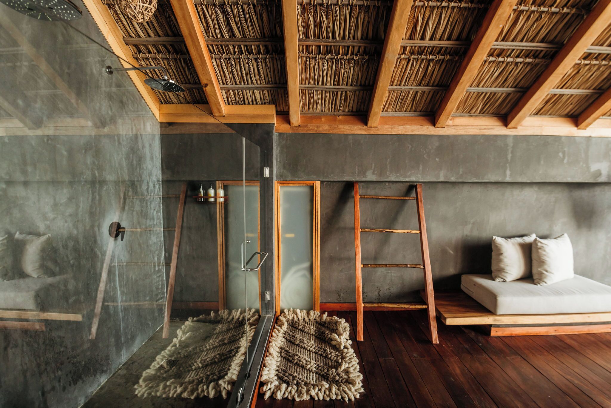 A modern room with a glass shower, wooden beams, a sofa, and a woven rug.