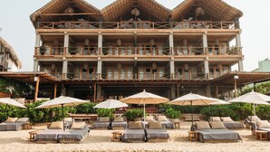 Un hôtel à plusieurs étages, doté d’une façade en bois, de balcons et d’un espace de détente en bord de plage, avec des chaises longues et des parasols.