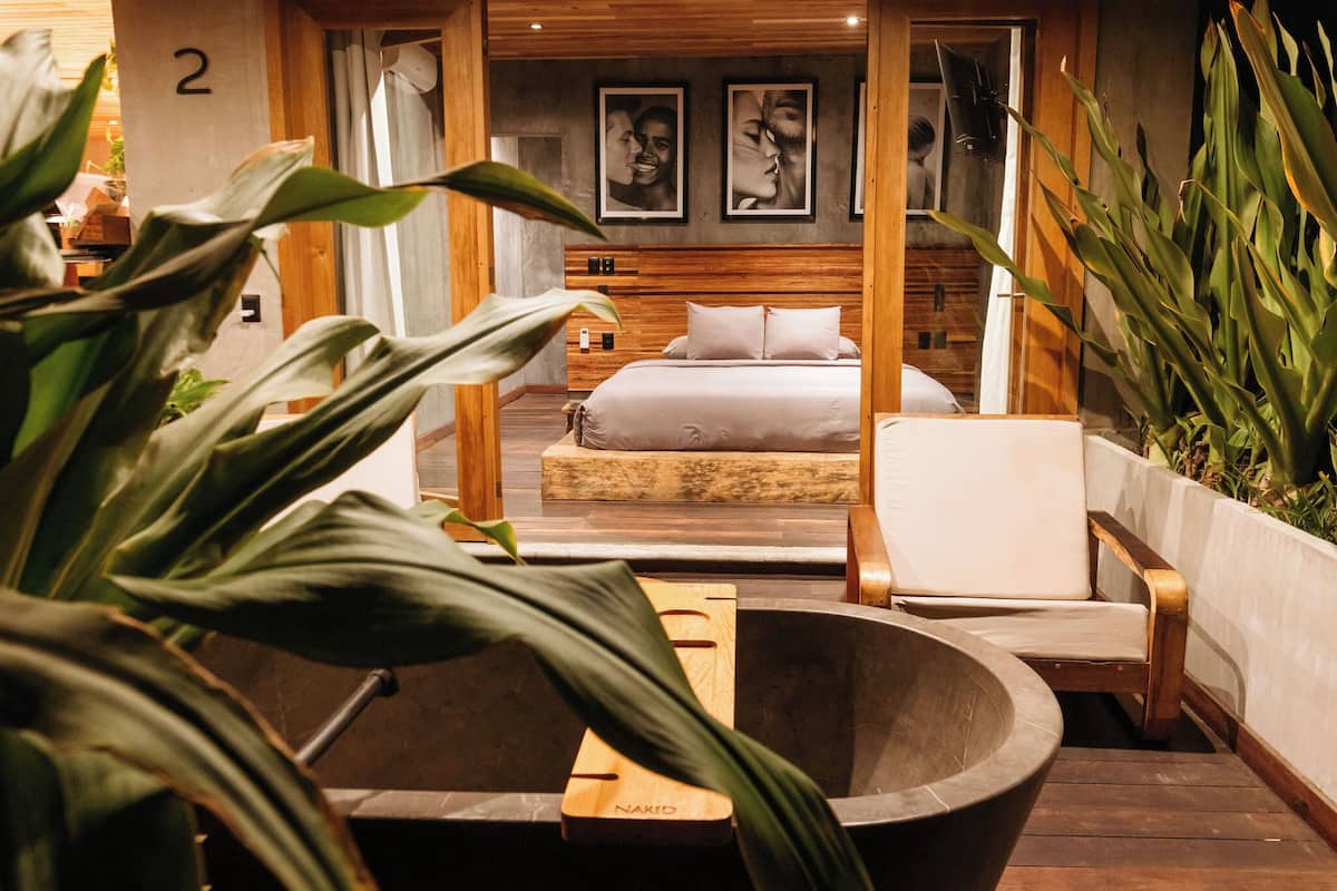 Een moderne slaapkamer met een groot bed, een houten hoofdbord en een houten bankje. Er staan planten en een grote kom op de voorgrond.