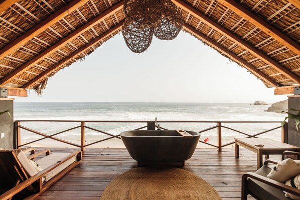 Eine Holzterrasse mit einer großen Badewanne, Liegestühlen und Blick auf das Meer.