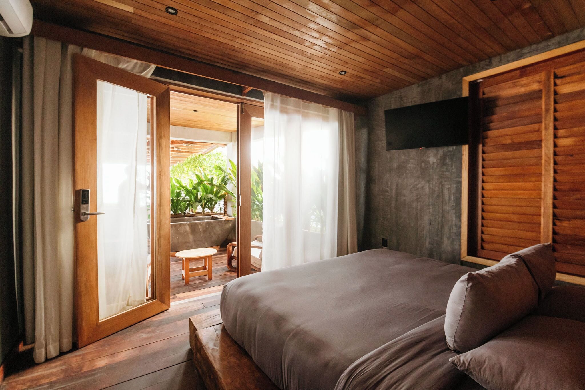 Een slaapkamer met een houten plafond, een bed met kussens, een aan de muur bevestigde televisie en een schuifdeur van glas die leidt naar een buitenruimte met planten.