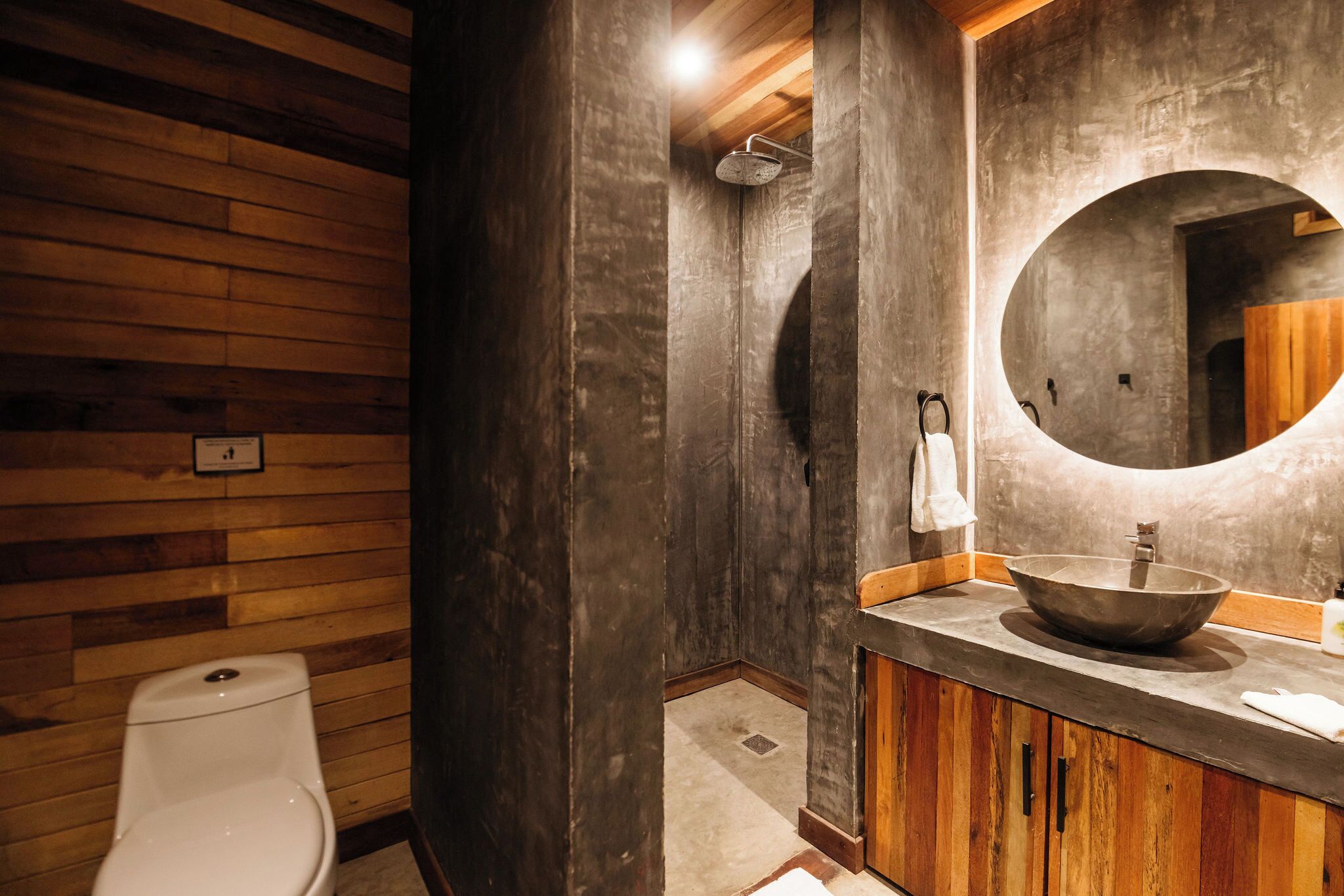 Un baño con pared de madera, un inodoro blanco, un espejo grande y un tocador de madera con lavabo.