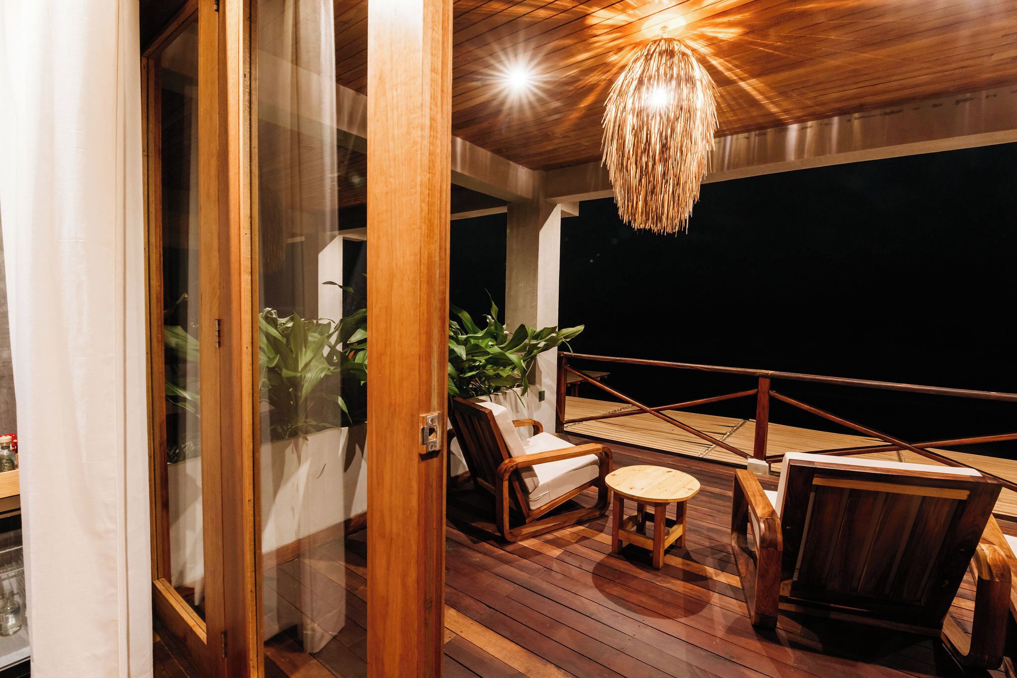 A wooden deck with a glass door leading to a room, a hanging light fixture, and a seating area with a small table and chairs.