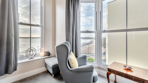 A room with a view of the sea, a grey armchair, a small table, and a window with curtains. - 3 bedroom accommodation in Tenby (Tenby)