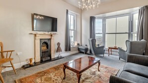 A spacious living room with a fireplace, a chandelier, a flat-screen TV, and large windows offering a view. - 3 bedroom accommodation in Tenby (Tenby)