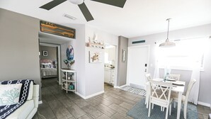 Ein geräumiger Wohnbereich mit Deckenventilator, einem Esstisch mit Stühlen und einer durchgehenden Tür erkennbaren Küche.