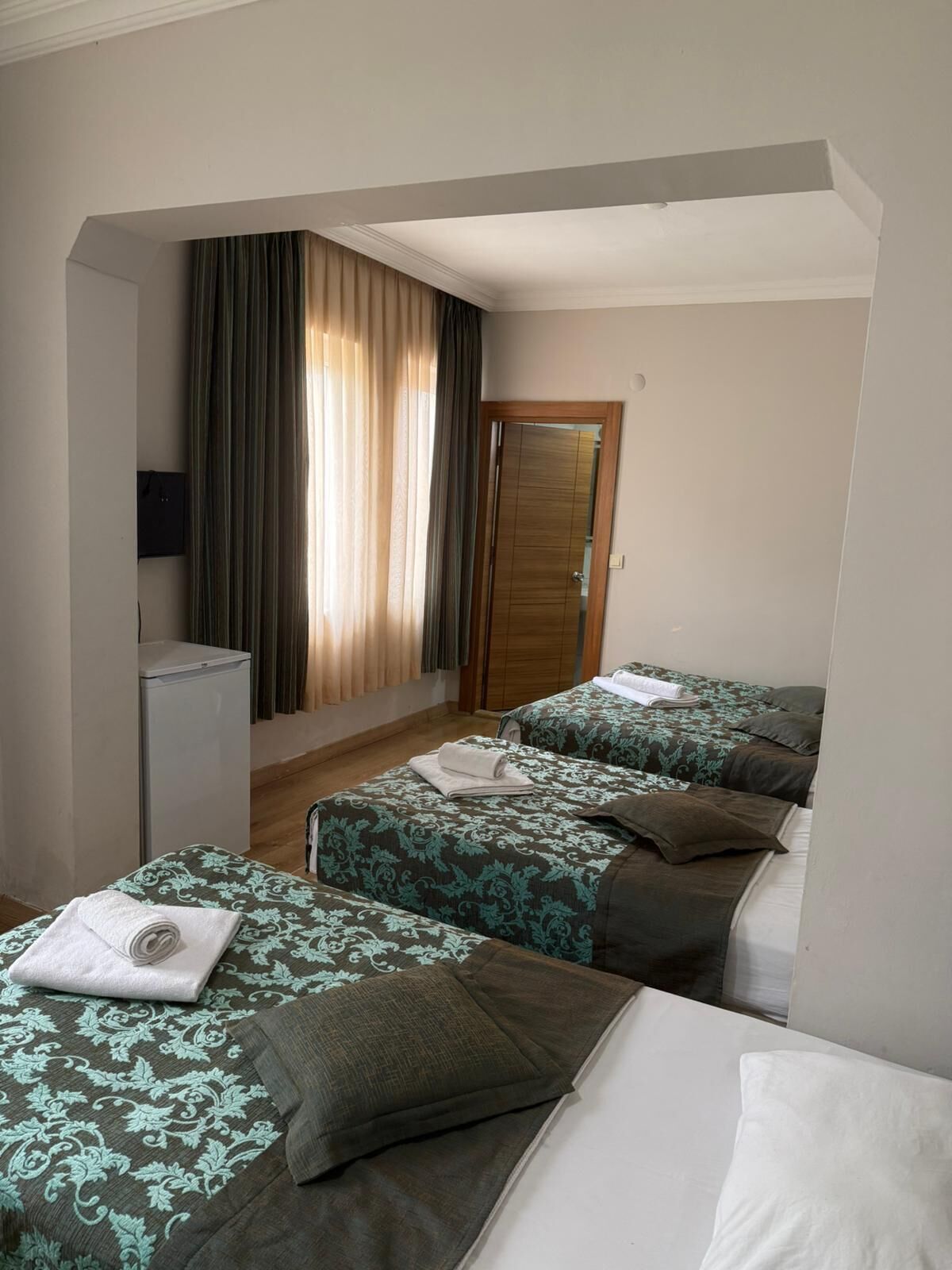 Ein Hotelzimmer mit zwei Einzelbetten, einem kleinen Kühlschrank, einer Holztür und einem Fenster mit Vorhängen.