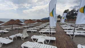 A beach with rows of white lounge chairs and thatched umbrellas, a pier in the background, and flags with 'Hotel Gold Stone' branding. - Hotel Gold Stone (Kemer)