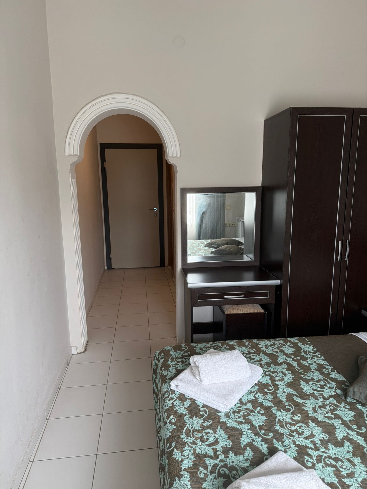 A hotel room with a bed, a desk with a television, a wardrobe, and a door leading to another room.