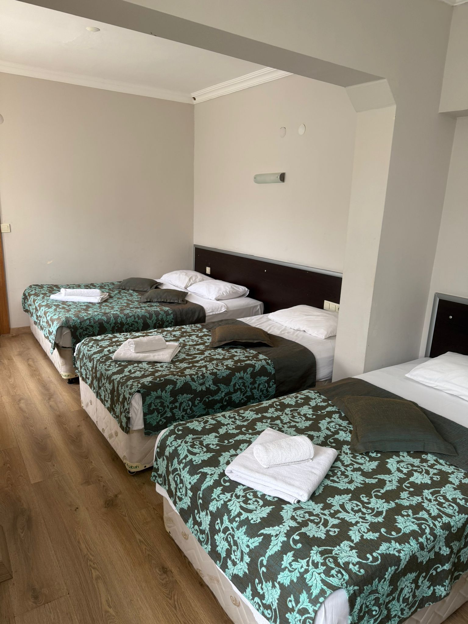 A hotel room with four single beds, each with a patterned bedspread and pillows, a bedside table, and a wall-mounted light fixture.