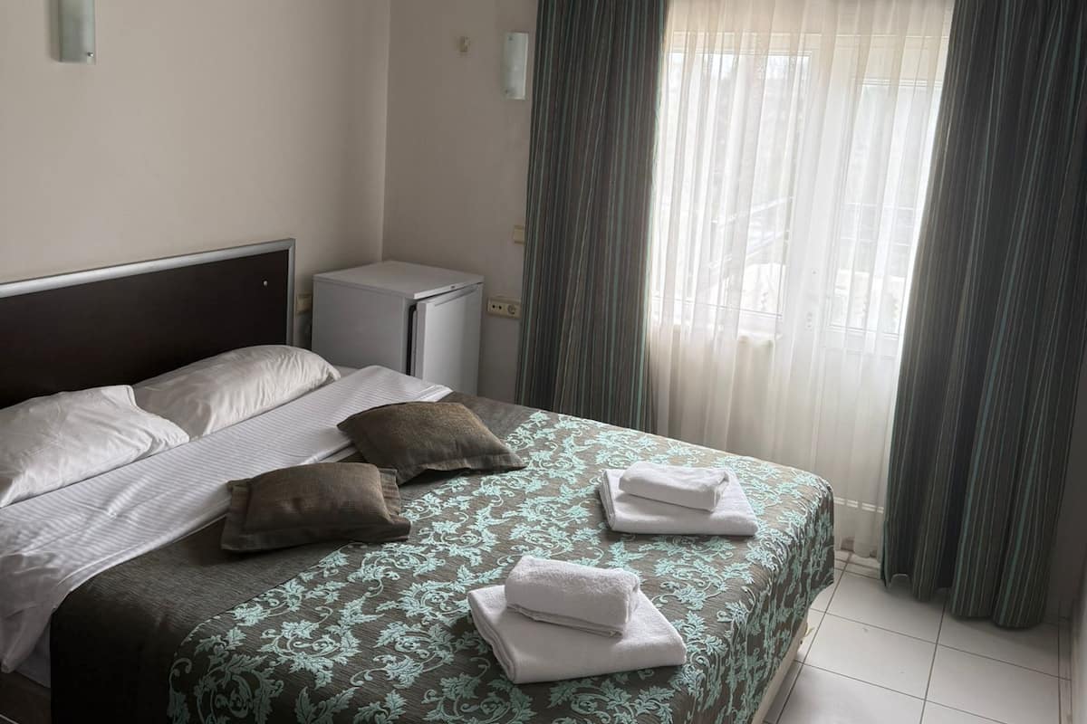 A hotel room with a bed, a desk, an air conditioner, a window with curtains, and a tiled floor.