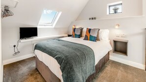 A bedroom with a sloped ceiling, a skylight, a bed with a green blanket, a TV on a shelf, and a small bedside table with a lamp. - We just love this romantic barn conversion. (Skirlaugh)