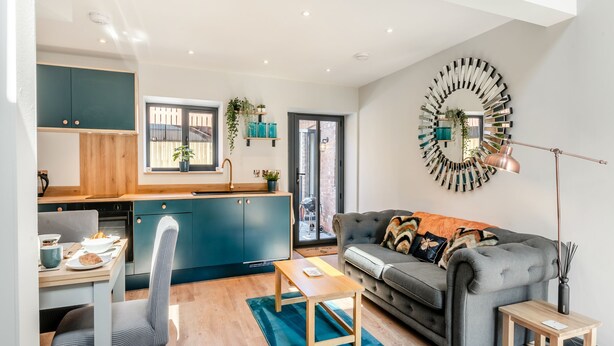 A modern kitchen with a window, a dining area with a table and chairs, a grey sofa with a patterned throw, and a unique wall clock. - We just love this romantic barn conversion. (Skirlaugh)