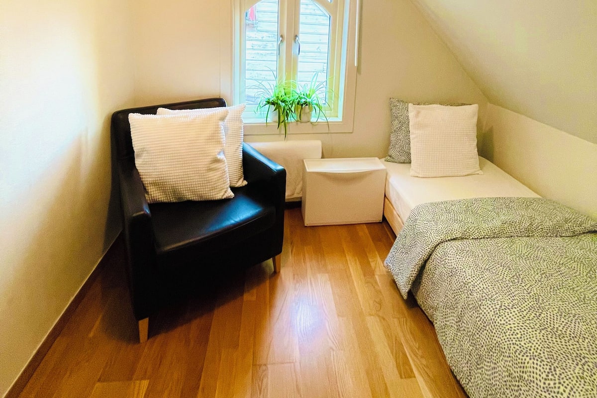 A small, cozy room with a black sofa, a small table, and a bed. There is a window with a view of the outside.